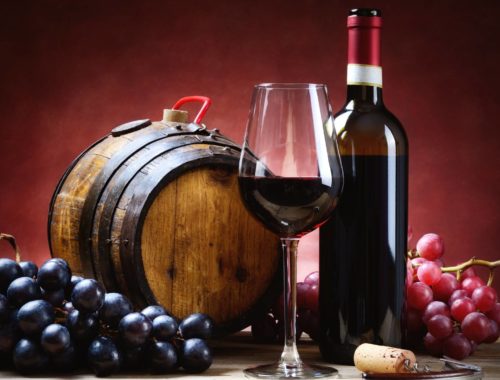 Grapes, berries, a wine glass (half full), a full bottle of wine, and keg are on a wooden table.